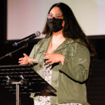 A woman wearing a mask speaking into a microphone.