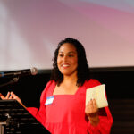 A woman standing at a podium holding a book and a microphone.