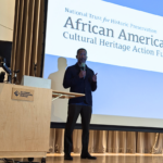 A person stands on stage, giving a thumbs up, with a presentation slide about the African American Cultural Heritage Action Fund in the background.