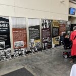 A person in a red coat speaks to someone in a mobility chair in front of display panels showcasing Oregon's civil rights history. The exhibit features images and text about struggles, resilience, and unity.