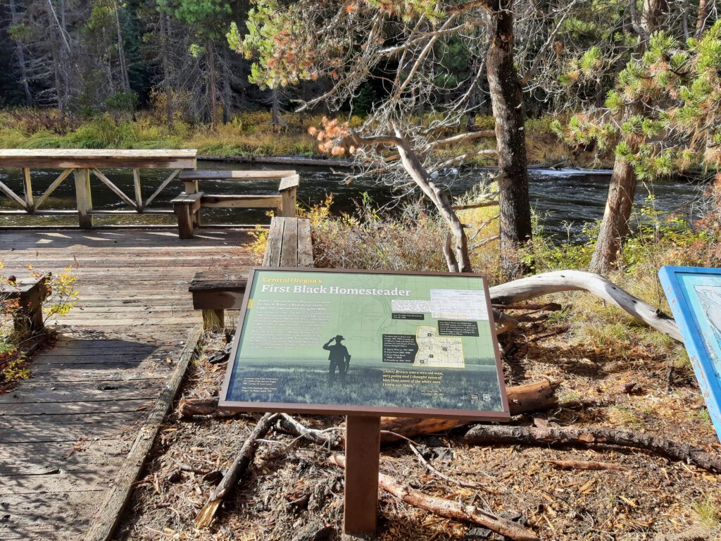 Information sign titled "First Black Homesteader" near a wooden deck by a river, surrounded by trees and autumn foliage.