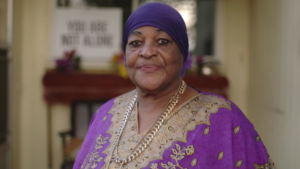 Smiling woman wearing a purple headscarf and tunic with gold patterns, standing indoors. A sign reading "You Are Not Alone" is blurred in the background.