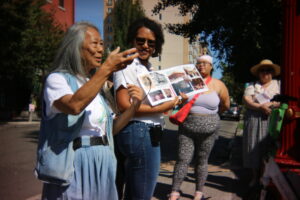 An older woman speaks while another person holds open a photo book; two other people stand nearby on a city street.