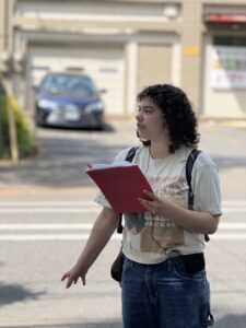A person with curly hair holds a red folder and gestures while standing outdoors near a street, wearing a t-shirt and jeans.
