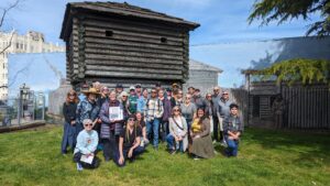 A group of people poses for a photo in front of a historic log structure and mural on a sunny day.