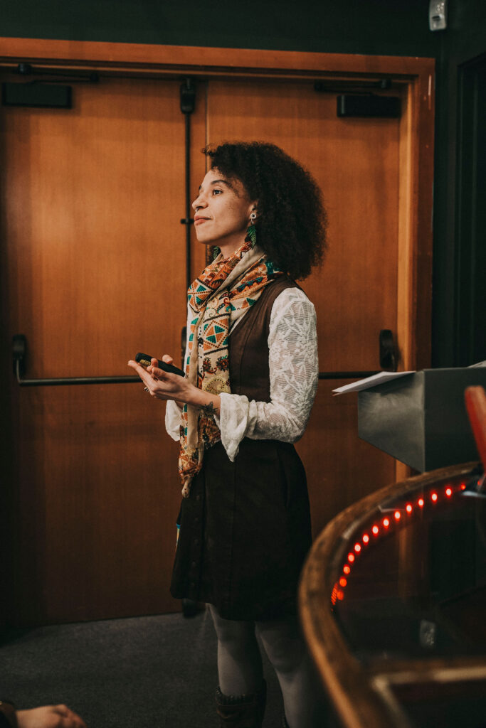 A woman stands indoors near a wooden door, wearing a patterned scarf, white lace blouse, and dark dress.