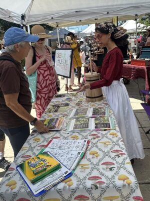 A woman in historical attire demonstrates butter churning at an outdoor market booth, while people observe and pamphlets