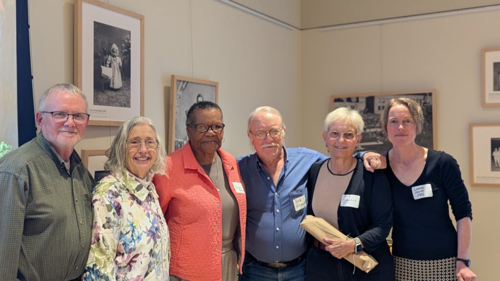 Six older adults stand together indoors, smiling for a group photo; some wear nametags. Framed black-and-white photos hang on the wall behind them