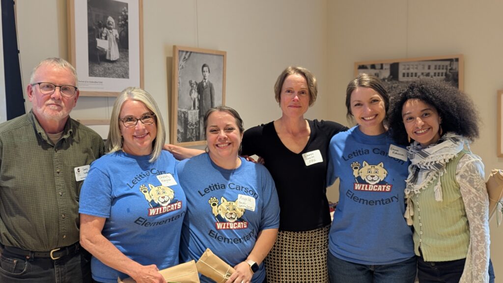 Six adults, three in matching Letitia Carson Elementary Wildcats shirts, stand together indoors, smiling at the camera. Framed photos are visible behind them