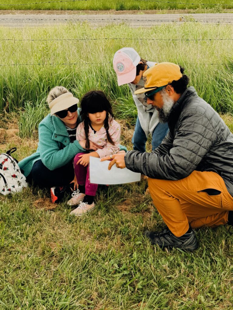 A man in a yellow cap shows a paper—perhaps the What is Letitia Carson Legacy Project News Newsletter?—to a woman and two children as they crouch together on grass near a fence, surrounded by tall greenery.