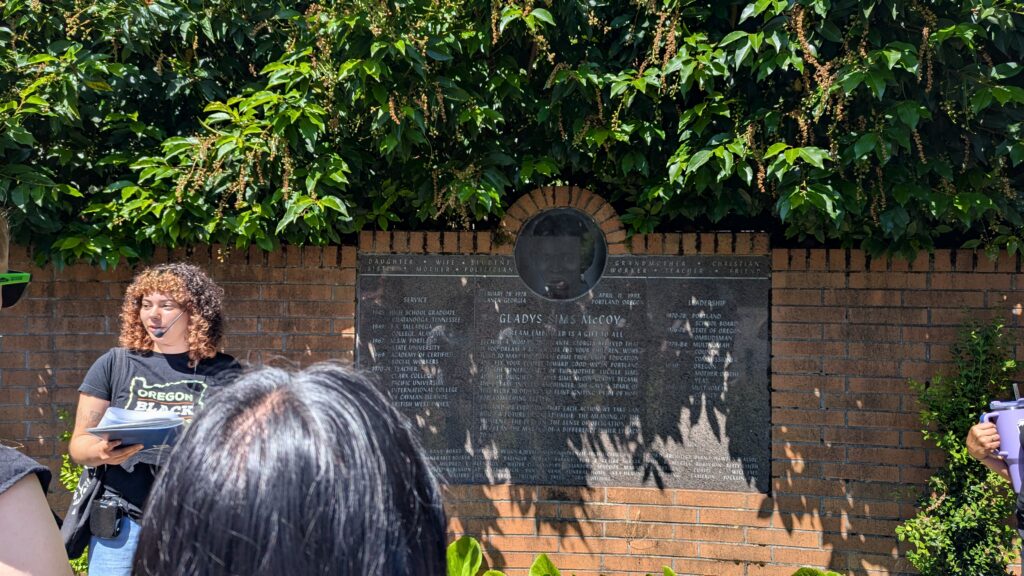 A group of people stands in front of a granite memorial plaque set against a brick wall, with greenery above and behind the plaque.