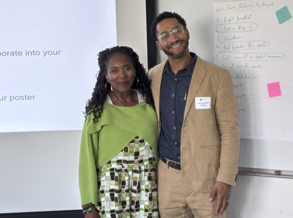 Two people stand side by side and smile in a classroom, with a whiteboard filled with handwritten notes visible in the background.