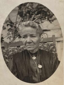 A vintage photo of Martha Lavadour with pulled back hair sits on a patterned chair outdoors, wearing a dark dress and two necklaces, looking slightly to the side.